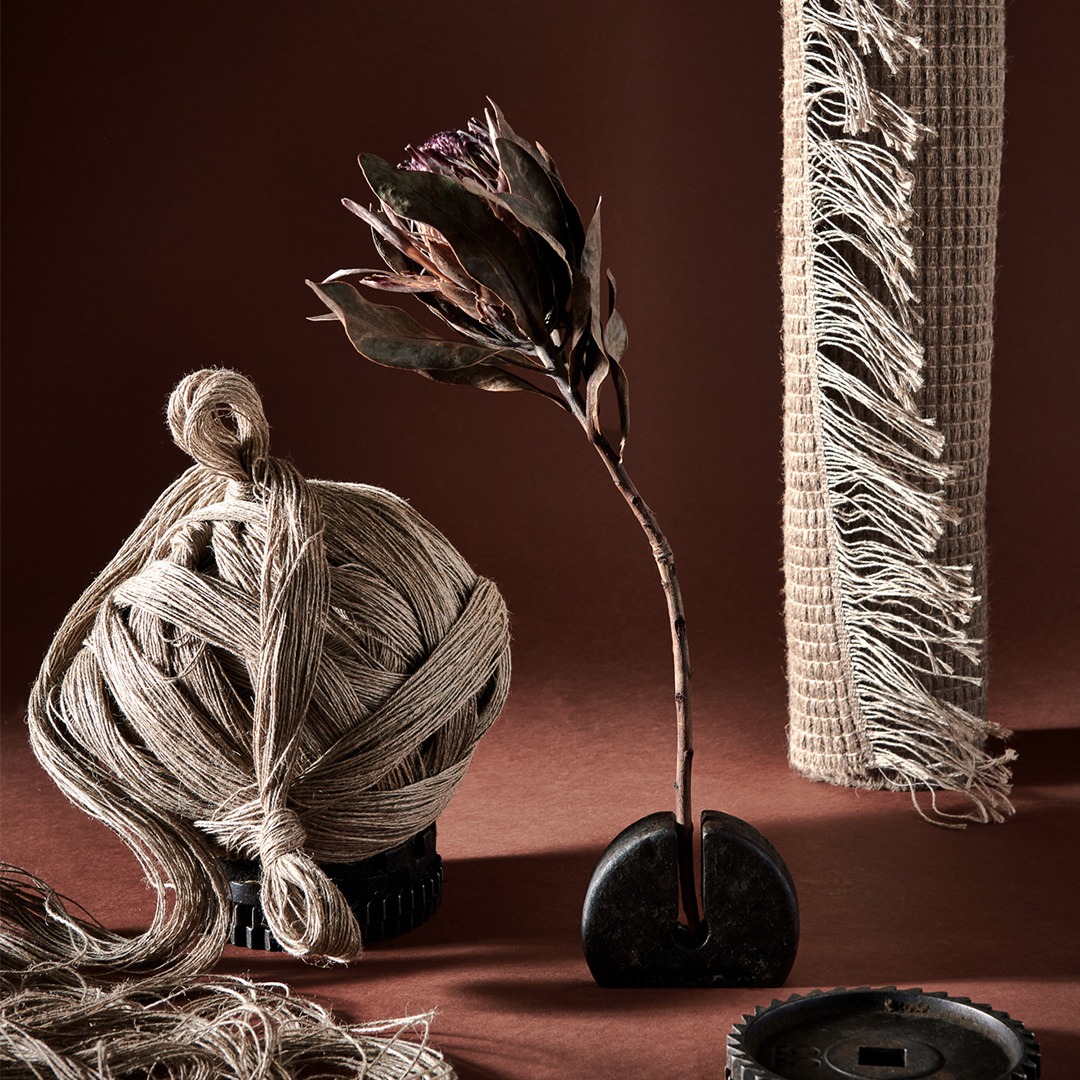 Une nature morte comprenant un tapis tissé en lin avec des franges, une pelote de fil de lin et une seule fleur séchée, le tout disposé sur une surface marron.